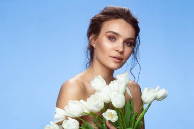Front portrait of the young girl with natural makeup and hairstyle looking at the camera, white tulips covered half of female's face, isolated over blue.