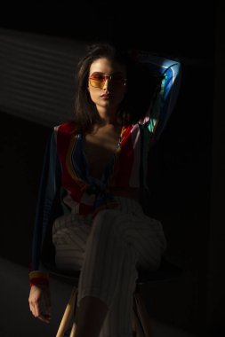 Renkli kıyafetler ve güneş gözlüğü takmış, boynun yakınında, koyu bir geçmişi olan genç ve şehvetli bir kadının ön portresi. Dikey görünüm.