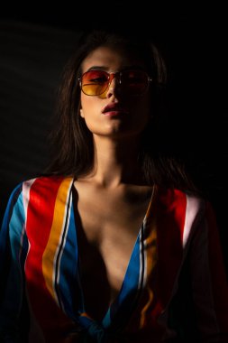 Renkli elbiseli, turuncu güneş gözlüklü, çekici bakışlı, izole edilmiş siyah arka planda genç, esmer bir kadının yakın plan görüntüsü..