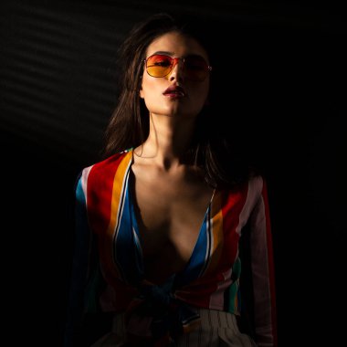Renkli bluzlu, güneş gözlüklü, koyu arkaplanlı genç bir kadının ön portresi. Dikey görünüm.