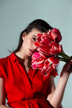 Genç esmer bir kadının ön görüntüsü kırmızı elbise giyiyor. Kırmızı lale demetinin yanında. Gri arka planda izole edilmiş kameraya bakıyor..