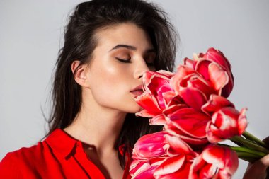 Kırmızı elbiseli çekici esmer kadın bir buket kırmızı lale kokusu alıyor, izole edilmiş gri arka plan..