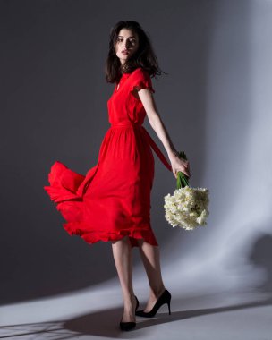 Uzun boylu, kırmızı elbiseli, elinde bir buket beyaz nergis tutan, izole edilmiş gri arka planlı esmer bir kadın..