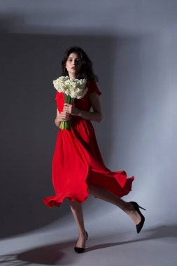 Kırmızı elbiseli esmer bir kadının, elinde bir buket beyaz nergis ve dönen gri bir arka planla tam boy görüntüsü..