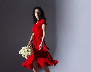 Kırmızı elbiseli esmer bir kadının elinde bir buket beyaz nergis ve dönen gri bir arka planla görüntüsü..