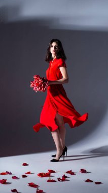 Kırmızı elbiseli, elinde bir buket lale tutan, poz veren ve kameraya bakan bir kadın resmi..
