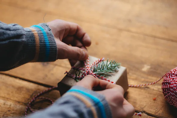 Christmas gift packaging. Men\'s hands in a blue sweater are packing a small gift box on a wooden table. Top view. Selective focus.