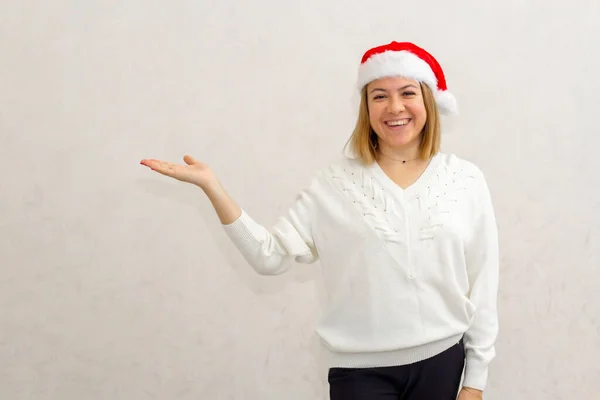 Vücut pozitif. Kısa saçlı, mutlu ve cüsseli bir kız Noel Baba 'nın Noel şapkasıyla ışıklı bir duvara yaslanmış, eli sağda duruyor. Boşluğu kopyala, düzeni kopyala.