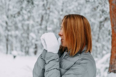 Gri ceketli, kısa saçlı ve beyaz eldivenli güzel bir kız karlı bir ormanda bir kupadan sıcak kahve içiyor. Kış mevsiminde sıcacık ve sıcak.