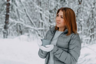 Gri ceketli, kısa saçlı ve beyaz eldivenli güzel bir kız karlı bir ormanda elinde bir kupa sıcak içecekle kendini ısıtıyor. Kış mevsiminde sıcacık ve sıcak.