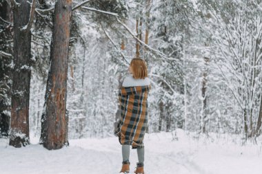 Ekose bir elbiseye sarılmış ve kış ormanında yürüyüş yapan mutlu bir genç kız. Noel tatilinde yürüyüş, seçici bir odak..