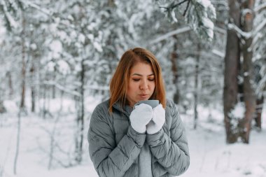 Gri ceketli ve beyaz eldivenli güzel bir kız karlı bir ormanda içki içiyor. Kış mevsiminde konfor ve sıcaklık. seçici odak.