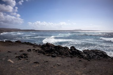 Koyu kahverengiden siyah lava dönüşen kayalar Atlantik Okyanusu 'nun batı kıyısını oluşturuyor. Derin turkuazdan mavi renge, rüzgarlı bir bahar gününde beyaz köpüklü dalgalarla. Lanzarote, Kanarya Adaları, İspanya.