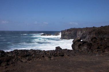 Koyu kahverengiden siyah lava dönüşen kayalar Atlantik Okyanusu 'nun batı kıyısını oluşturuyor. Derin turkuazdan mavi renge, rüzgarlı bir bahar gününde beyaz köpüklü dalgalarla. Lanzarote, Kanarya Adaları, İspanya.