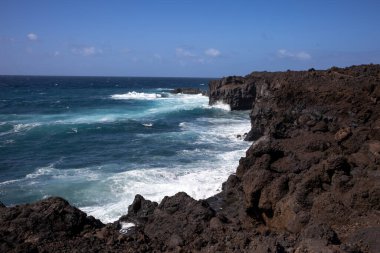 Koyu kahverengiden siyah lava dönüşen kayalar Atlantik Okyanusu 'nun batı kıyısını oluşturuyor. Derin turkuazdan mavi renge, rüzgarlı bir bahar gününde beyaz köpüklü dalgalarla. Lanzarote, Kanarya Adaları, İspanya.