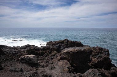Dark brown to black color of the lava stones and rocks, created a cliff above the wild water of the Atlantic Ocean. Cloudy sky in the early spring. Horizon of the ocean. Los Hervideros, Lanzarote, Canarian Islands, Spain.