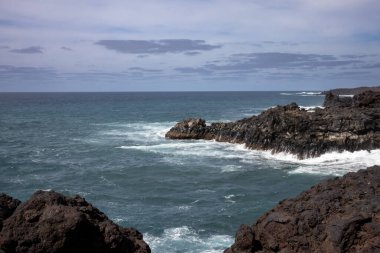 Dark brown to black color of the lava stones and rocks, created a cliff above the wild water of the Atlantic Ocean. Cloudy sky in the early spring. Horizon of the ocean. Los Hervideros, Lanzarote, Canarian Islands, Spain.