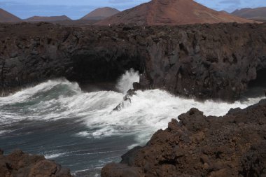 Dark brown to black color of the lava stones and rocks, created a cliff above the wild water of the Atlantic Ocean. Cloudy sky in the early spring. Mountains in the background. Los Hervideros, Lanzarote, Canarian Islands, Spain.