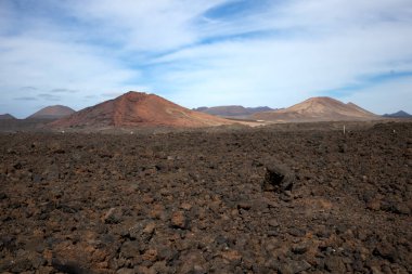 Lava soil of a brown to black color. Mountains in the background. Blue sky with white clouds in the early spring. Lanzarote, Las Palmas, Canary Islands, Spain.