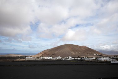 Black dry soil and village created by white houses of an unified local style. Hill in the background. Horizon of Atlantic Ocean. Cloudy sky. Lanzarote, Canary Islands, Spain.