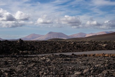 National Park of the volcanic formations. Colors of the land from red and brown to black. Just a road with limited traffic to preserve the heritage. Blue sky with white clouds. Timanfaya, Lanzarote, Canary Islands, Spain.
