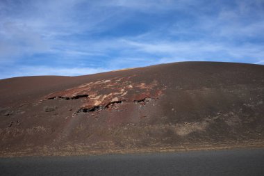 National Park of the volcanic formations. Colors of the land from red and brown to black. Just a road with limited traffic to preserve the heritage. Blue sky with white clouds. Timanfaya, Lanzarote, Canary Islands, Spain.