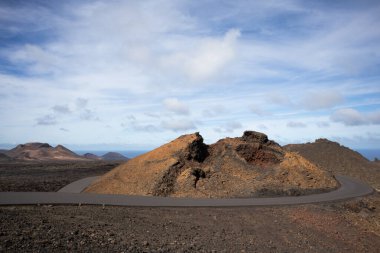 National Park of the volcanic formations. Colors of the land from red and brown to black. Just a road with limited traffic to preserve the heritage. Blue sky with white clouds. Timanfaya, Lanzarote, Canary Islands, Spain.