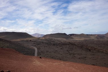 National Park of the volcanic formations. Colors of the land from red and brown to black. Just a road with limited traffic to preserve the heritage. Blue sky with white clouds. Timanfaya, Lanzarote, Canary Islands, Spain.