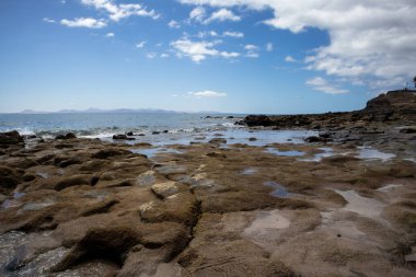 Lanzarote 'nin güneyindeki Rocky Sahili. Atlantik okyanusunun sakin suları. Fuerteventura adası ufukta. Beyaz bulutlu mavi gökyüzü. Playa Blanca, Lanzarote, Kanarya Adaları, İspanya.
