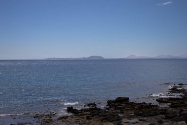 Lanzarote 'nin güneyindeki Rocky Sahili. Atlantik okyanusunun sakin suları. Fuerteventura adası ufukta. Beyaz bulutlu mavi gökyüzü. Playa Blanca, Lanzarote, Kanarya Adaları, İspanya.