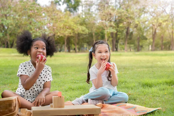 Happy cheerful white girls and black girl eat apple together at outdoors park , Relationship little kids, Diverse ethnic concept.