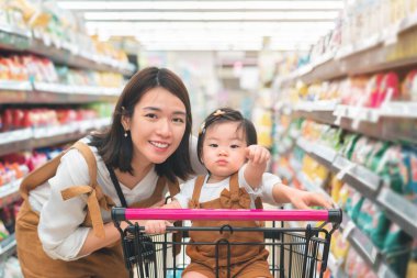 Asyalı anne ve kızı büyük bir süpermarketten yiyecek alıyorlar, Bebek tramvayda oturuyor, aile alışverişi konsepti.
