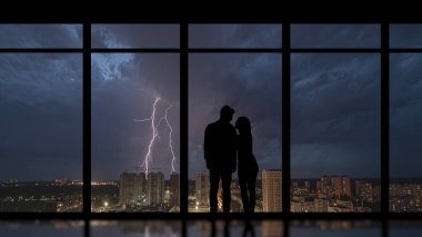 Gece yıldırımına karşı panoramik bir pencerenin yanında duran adam ve kadın.