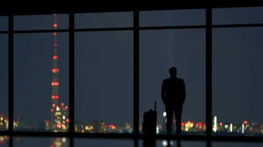 Şehrin arka planında panoramik pencerenin yanında duran bavullu adam.