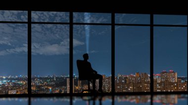 Gece şehir arka planında panoramik pencerenin yanında oturan erkek.