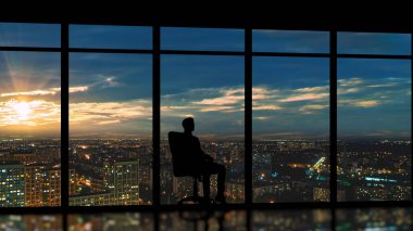 Erkek, şehrin arka planında panoramik pencerenin yanında oturuyor.