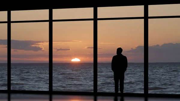 The man standing near the panoramic window against the sea sunset