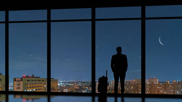 那个提着手提箱站在一座夜城的全景窗前的男人 — 图库照片