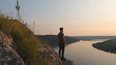 The young man stands on rocky mountain on beautiful river background