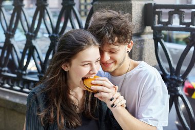 Genç bir adam ve bir kız ellerinde hamburgerler tutuyor. Adam kıza hamburgerinden bir ısırık veriyor..