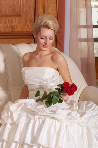 Indoor portrait of sexy bride with blonde hair sitting on a sofa, holding red roses. — Stock Photo, Image