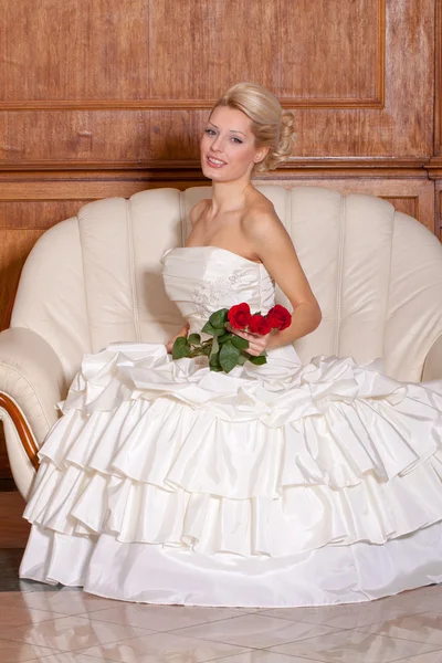 Indoor portrait of sexy bride with blonde hair sitting on a sofa, holding red roses. — Stock Photo, Image