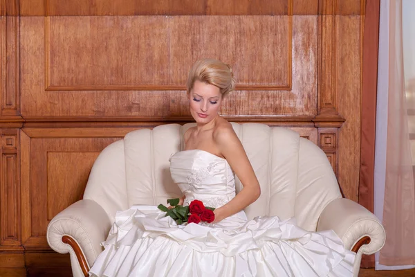 Portrait intérieur d'une belle mariée aux cheveux blonds assise sur un canapé, tenant des roses rouges . — Photo