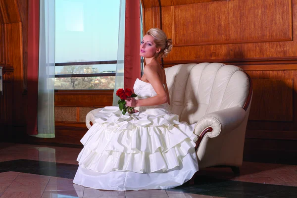 Indoor portrait of adorable bride with blonde hair sitting on a sofa, holding red roses. — Stock Photo, Image