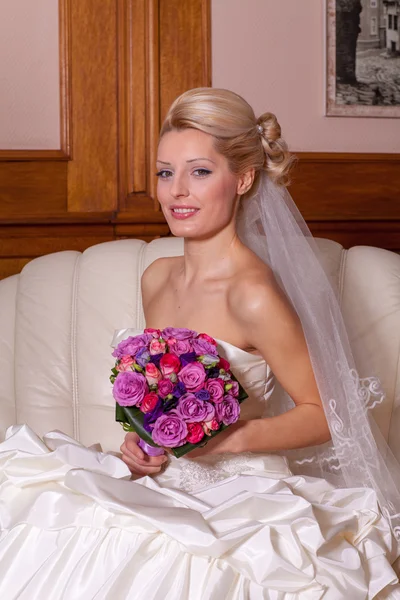 Indoor portrait of sexy bride with blonde hair sitting on a sofa. — Stock Photo, Image