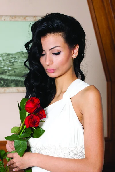 Indoor portrait of adorable bride with black long hair holding red roses. — Stock Photo, Image