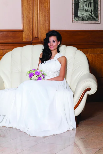 Indoor portrait of sexy bride with black long hair sitting on a sofa. — Stock Photo, Image