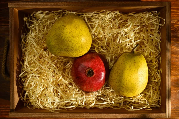 Dvě zralá hrušky a šťavnaté granátové jablko v krabici s hoblinami na dřevěném stole, detailní pohled shora — Stock fotografie