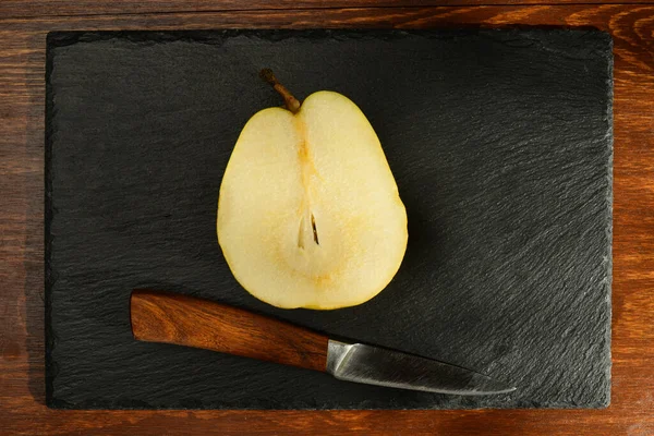 Half natural yellow pear and knife on a slate board, close-up, on a wooden table, top view — Stock Photo, Image