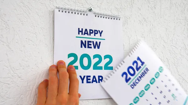 2021 'in Aralık sayfasındaki duvar takviminin yırtıldığı erkek ellerinin yakın çekimi. Ardından yeni bir 2022 takviminin başlık sayfası.